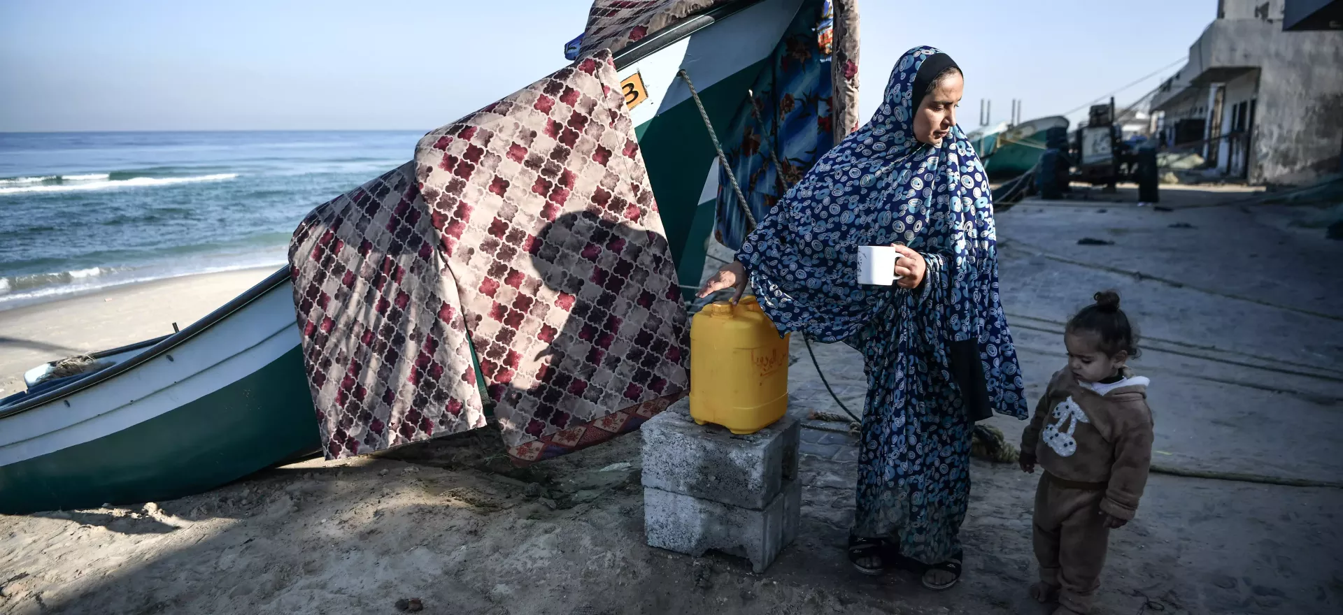 Una madre y su hija pequeña descansan y beben agua junto a un barco de pesca en la playa.