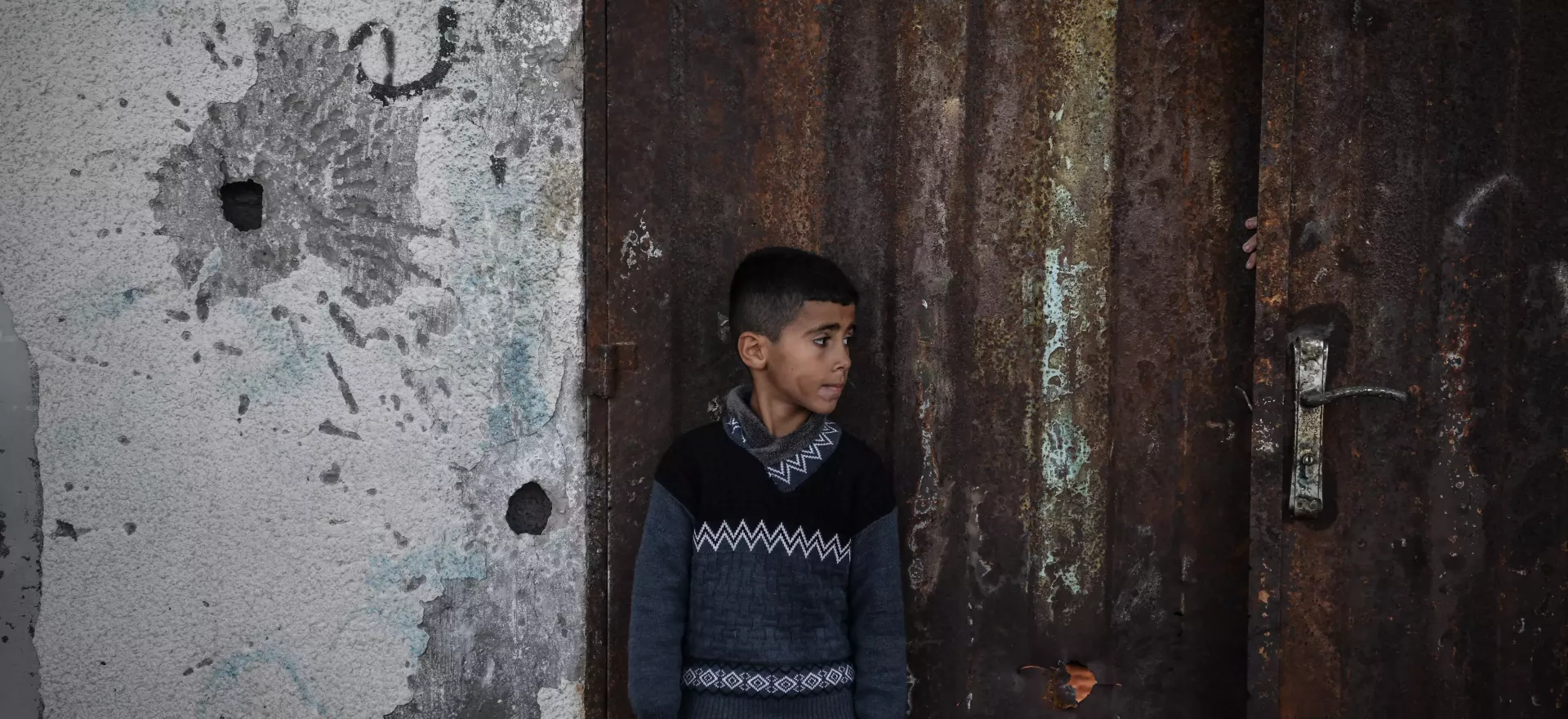 A boy stands next to a bullet ridden wall and a rusted door.