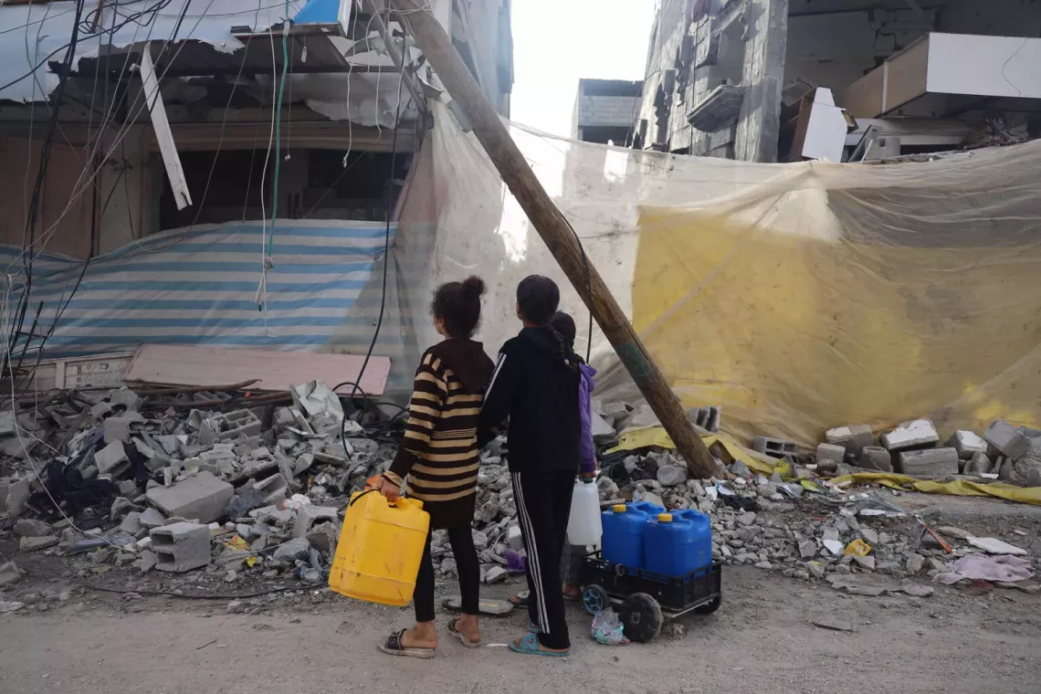 أطفال يجلبون الماء يمرون بجانب مبانٍ متضررة بشدة في قطاع غزة.