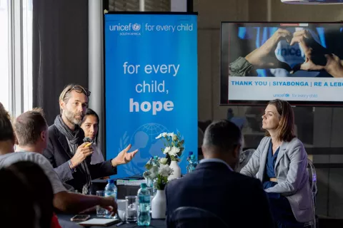 The UNICEF Communications chief speaks while holding a microphone in his hand with executives around him listening with interst.