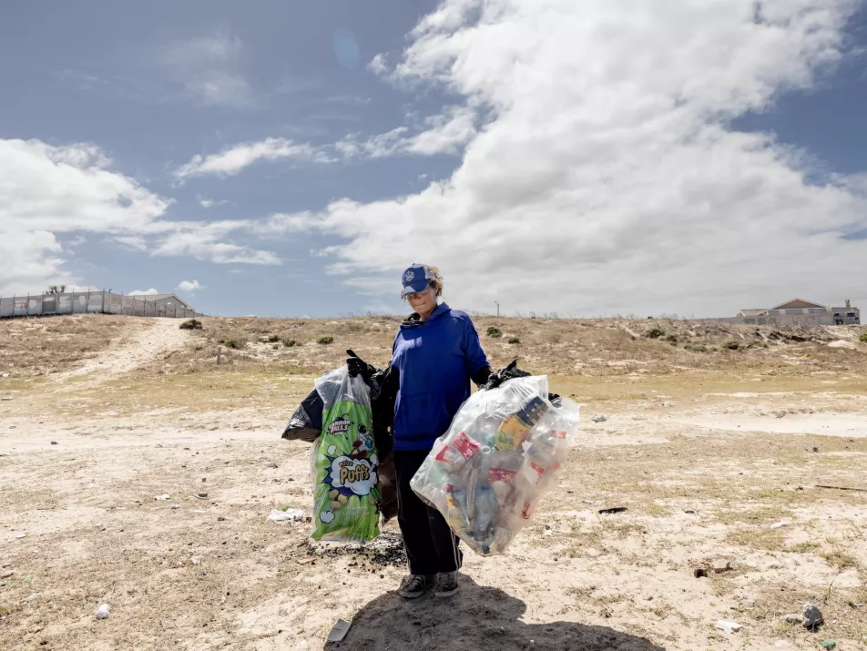 A woman carrying large bags of waste she's collected, walks across a bare stretch of land.