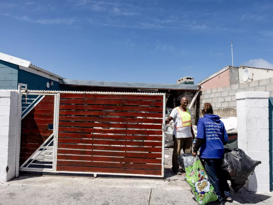 A woman carries bags of collected recyclables up to a gate where a man is waiting for her.