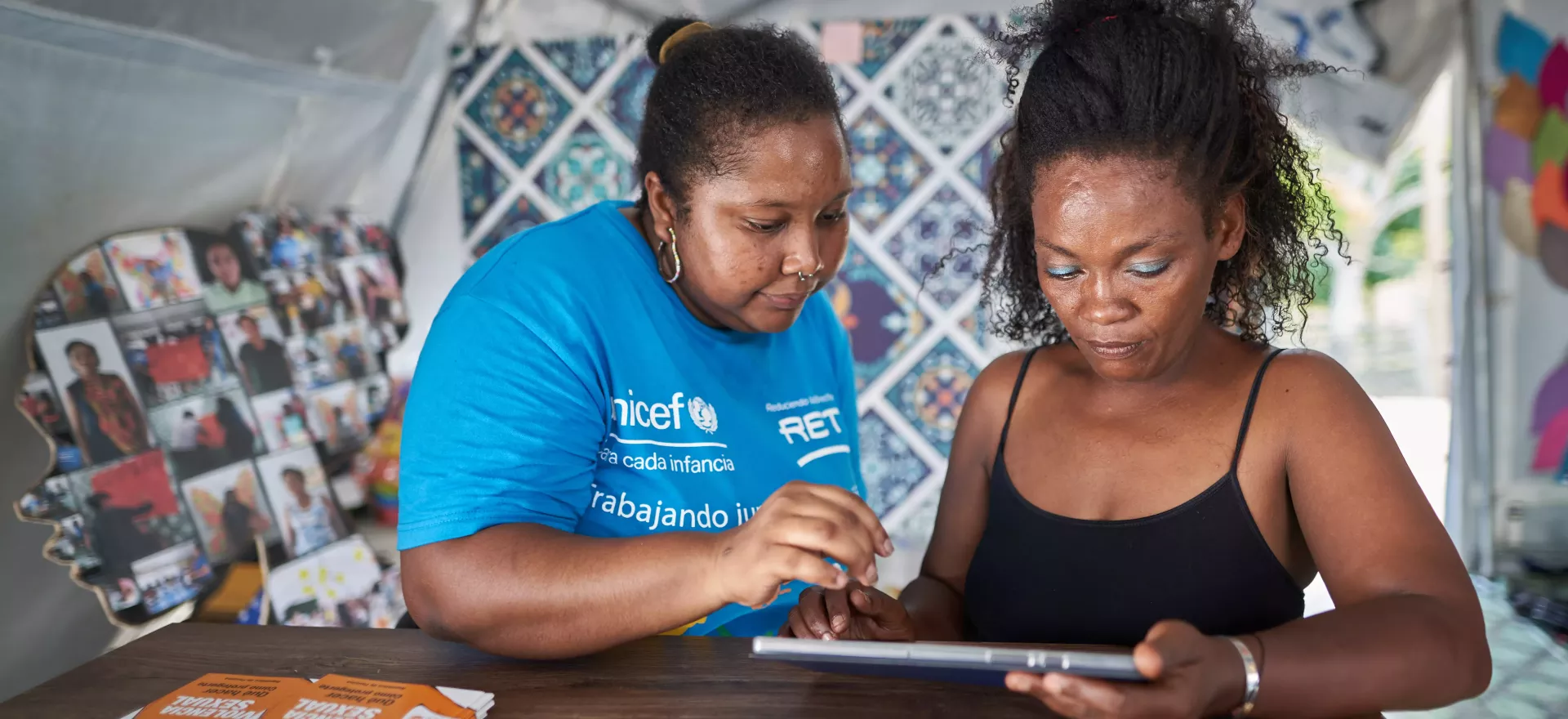 一名身穿联合国儿童基金会衬衫的妇女帮助另一名妇女使用平板电脑。
