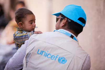 De dos, un homme avec une veste UNICEF, porte une petite fille dans ses bras