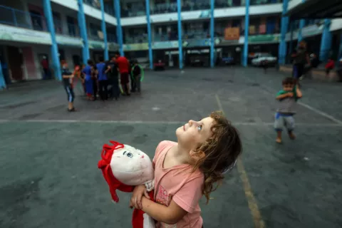 Aya, a 5-year-old girl clutching her doll to ease her fear, gazing at Gaza's sky filled with warplanes from inside an UNRWA school in the Gaza Strip.