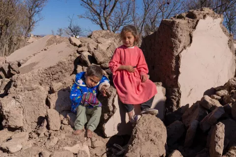 children sit on a pile of rubble where their home used to stand.