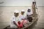 3名穿着白色T恤、戴着帽子的孩子坐在一条船上，一位成年人正在划船。