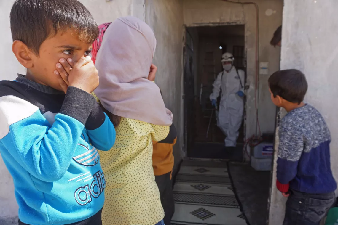 Children in Syria cover their mouths and noses as a member of the Syrian Civil Defence disinfects a former school building, now inhabited by displaced families, to prevent the spread of COVID-19.