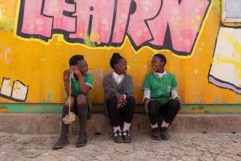 Three adolescent girls in green school uniforms sit on a small ledge in front of a yellow wall with graffiti painted on it.