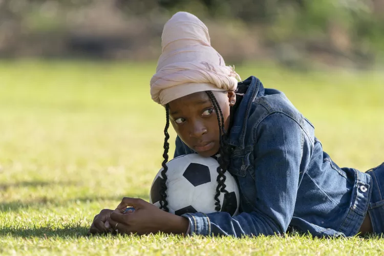On a sport field, a girl with her chin resting on a football looks into the distance.
